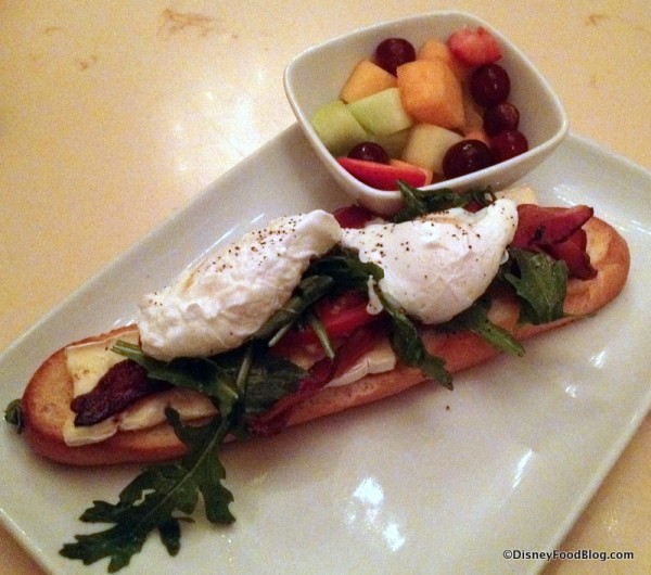 Open-Face Bacon and Egg Sandwich with poached eggs, applewood bacon, and brie cheese on a toasted baguette served with fresh fruit